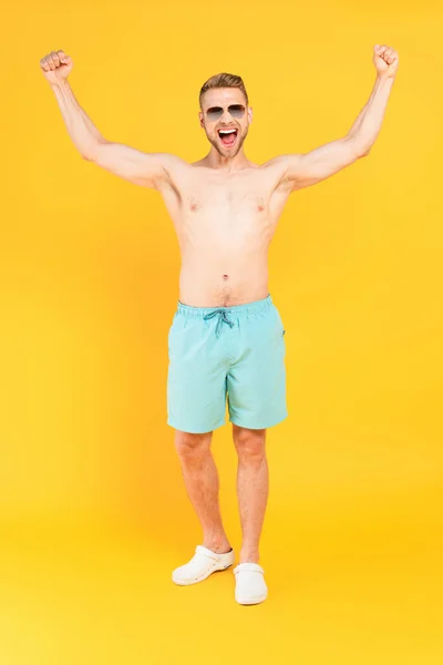 Homme excité et torse nu dans des lunettes de soleil debout avec les mains au-dessus de la tête sur jaune — Photo de stock