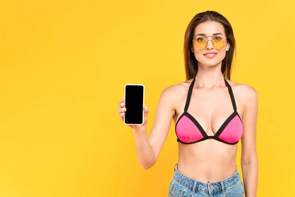 Весёлая девушка в солнечных очках держит смартфон с пустым экраном, изолированным на жёлтом — стоковое фото