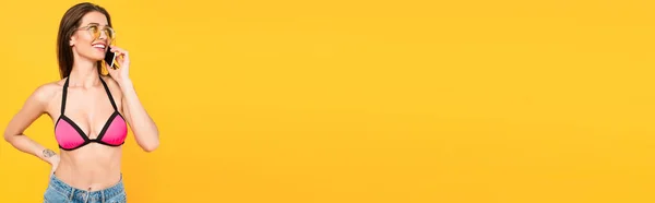 Plano panorámico de chica feliz en traje de baño y gafas de sol hablando en el teléfono inteligente aislado en amarillo - foto de stock