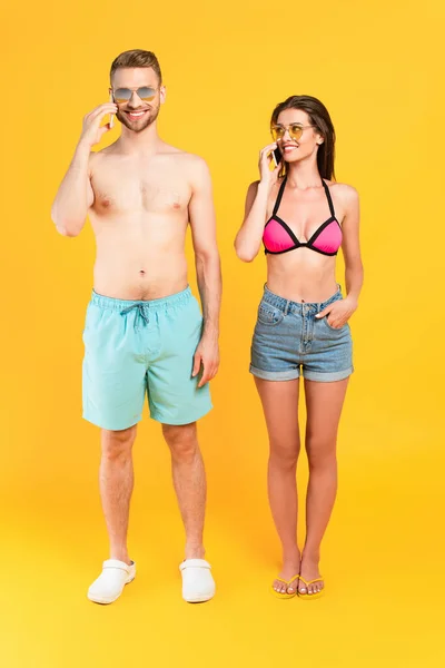 Щаслива пара в сонцезахисних окулярах і купальниках розмовляє на смартфонах на жовтому — стокове фото