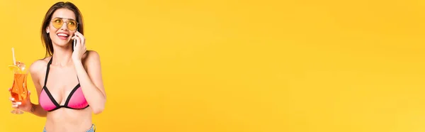 Plan panoramique de femme heureuse en lunettes de soleil parlant sur smartphone et tenant cocktail isolé sur jaune — Photo de stock