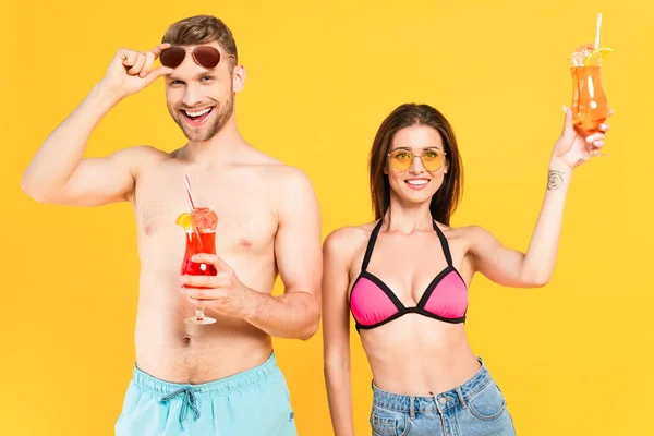 Веселая пара в солнцезащитных очках и купальниках с коктейлями, изолированными на желтом — стоковое фото