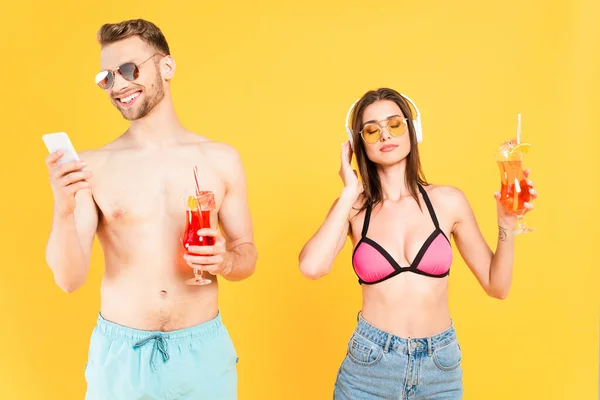 Hombre feliz en gafas de sol utilizando el teléfono inteligente y la celebración de cóctel, mientras que la chica en traje de baño escuchar música aislada en amarillo - foto de stock