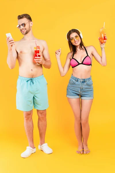 Happy man in sunglasses using smartphone and holding cocktail while girl in swimwear listening music on yellow — Stock Photo
