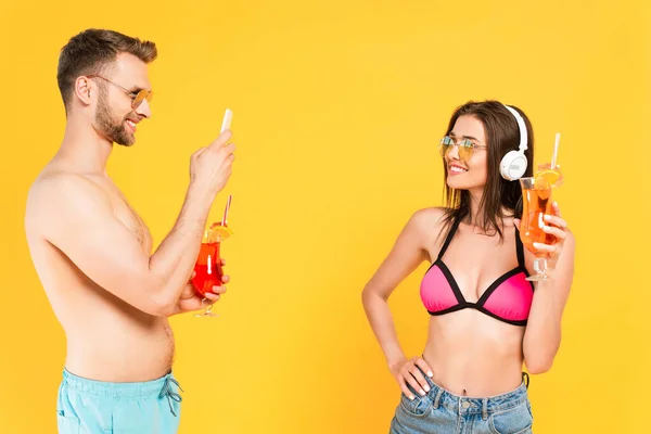 Hombre barbudo tomando fotos de chica feliz en auriculares con cóctel aislado en amarillo - foto de stock