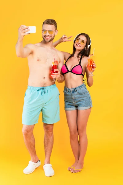 Happy girl in headphones showing peace sign and taking selfie with man holding cocktail on yellow — Stock Photo