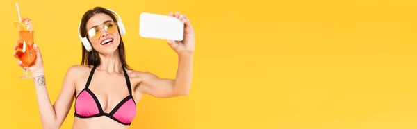Concept panoramique de fille joyeuse et tatouée dans des lunettes de soleil prendre selfie tout en tenant cocktail isolé sur jaune — Photo de stock
