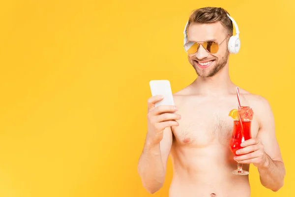 Cheerful and shirtless man in sunglasses using smartphone while holding cocktail isolated on yellow — Stock Photo