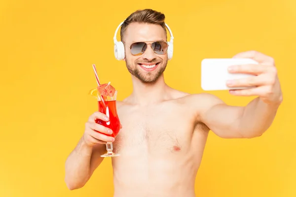 Messa a fuoco selettiva di uomo felice e senza maglietta in occhiali da sole e cuffie prendere selfie mentre tiene cocktail isolato su giallo — Foto stock