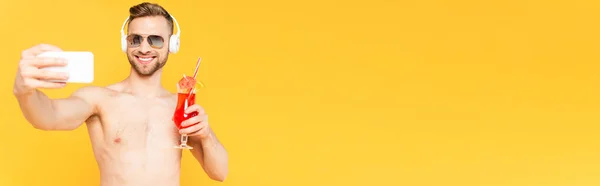 Image horizontale d'un homme torse nu portant des lunettes de soleil et des écouteurs prenant du selfie tout en tenant un cocktail isolé sur du jaune — Photo de stock