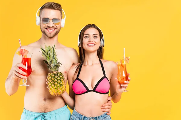 Homme et femme heureux dans des écouteurs tenant l'ananas et des cocktails isolés sur jaune — Photo de stock