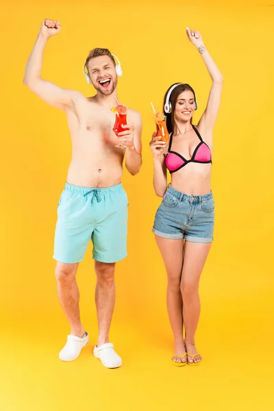 Feliz hombre y mujer en auriculares con cócteles y de pie en amarillo - foto de stock