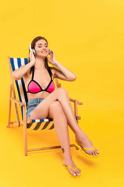 Mujer feliz en traje de baño y auriculares sentados en la silla de cubierta mientras escucha música en amarillo - foto de stock