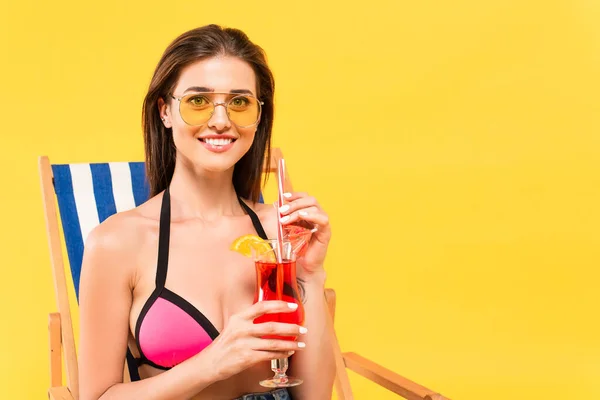 Felice donna in occhiali da sole in possesso di cocktail mentre seduto sulla sedia a sdraio isolato su giallo — Foto stock