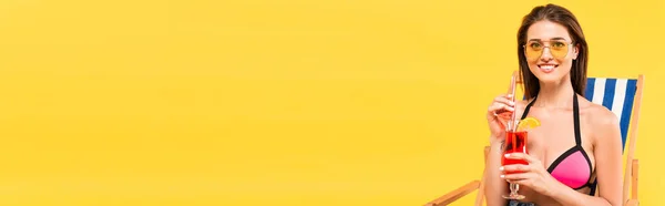 Raccolto panoramico di donna felice in occhiali da sole in possesso di cocktail mentre seduto sulla sedia a sdraio isolato su giallo — Foto stock