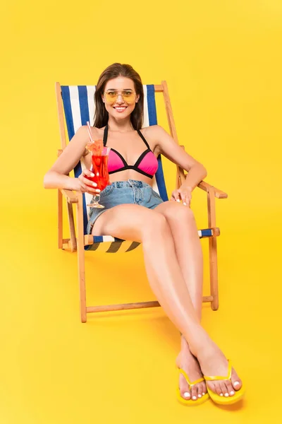 Cheerful woman in sunglasses and flip flops sitting on deck chair and holding cocktail on yellow — Stock Photo