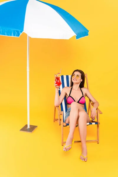 Весела жінка в сонцезахисних окулярах сидить на стільці і тримає коктейль біля пляжної парасольки на жовтому — стокове фото