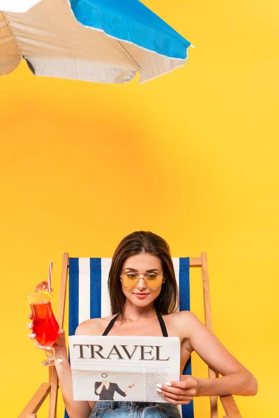 Schöne Frau sitzt auf Liegestuhl mit Cocktail beim Lesen Reisezeitung in der Nähe Sonnenschirm auf gelb — Stockfoto