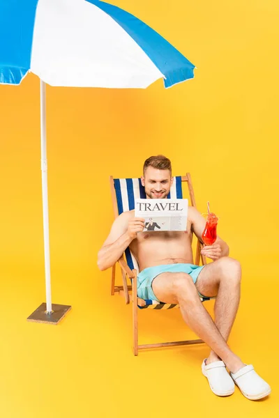 Handsome man sitting on deck chair with cocktail while reading travel newspaper near beach umbrella on yellow — Stock Photo