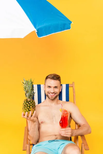 Happy shirtless man sitting on deck chair with cocktail and pineapple near beach umbrella on yellow — Stock Photo