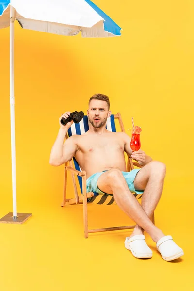 Surprised shirtless man holding cocktail and binoculars while sitting on deck chair on yellow — Stock Photo