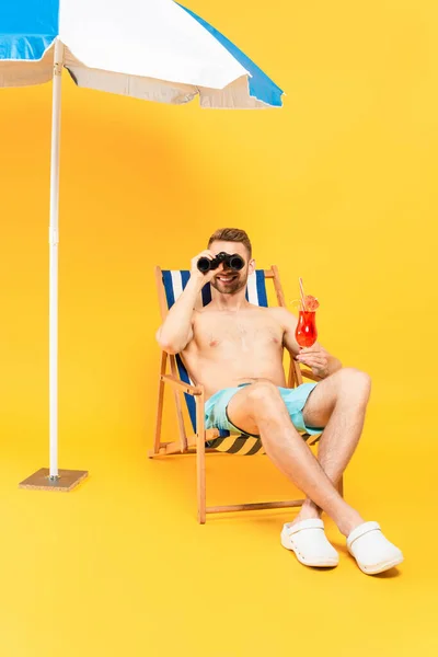 Cheerful shirtless man holding cocktail and looking through binoculars while sitting on deck chair on yellow — Stock Photo