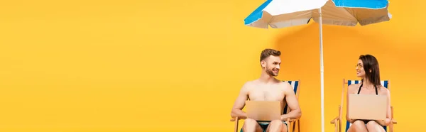 Panoramic shot of couple with laptops sitting on deck chairs near beach umbrella on yellow — Stock Photo