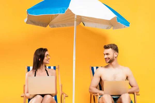 Happy couple with laptops sitting on deck chairs near beach umbrella on yellow — Stock Photo