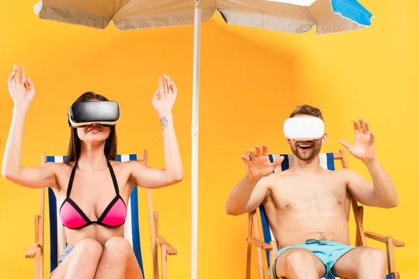 Smiling couple in virtual reality headsets sitting on deck chairs and gesturing near beach umbrella on yellow — Stock Photo