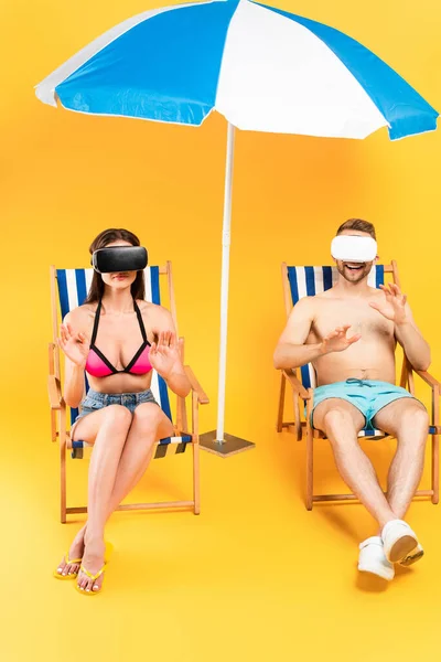 Щаслива пара у гарнітурах віртуальної реальності, що сидять на стільцях палуби і жестикулюють біля пляжної парасольки на жовтому — стокове фото