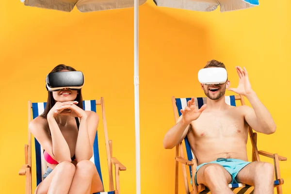 Happy couple in virtual reality headsets sitting on deck chairs near beach umbrella on yellow — Stock Photo