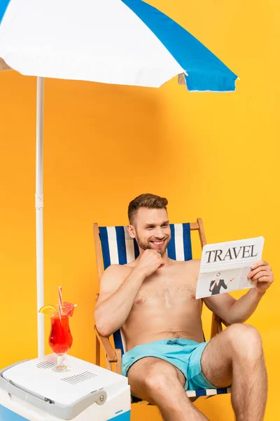 Shirtless man smiling while reading travel newspaper and sitting on deck chair near cocktail on portable fridge freezer on yellow — Stock Photo