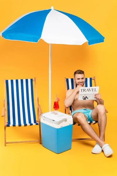 Muscular man smiling while reading travel newspaper and sitting on deck chair near cocktail on portable fridge freezer on yellow — Stock Photo