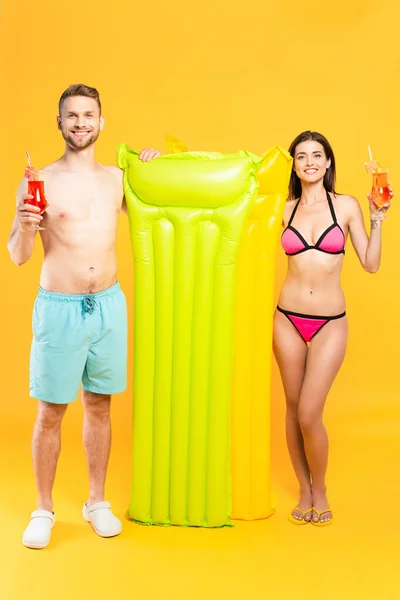 Счастливая пара в купальниках смотрит в камеру и держит коктейли возле надувного матраса на желтом — стоковое фото