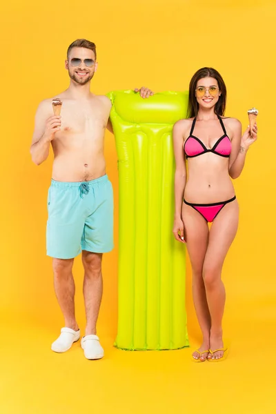 Homme musclé heureux et femme gaie en maillots de bain et lunettes de soleil tenant la crème glacée tout en se tenant près du matelas gonflable sur jaune — Photo de stock