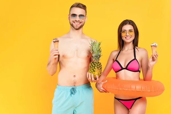 Счастливый мускулистый мужчина с ананасом рядом с женщиной в солнечных очках, держащий мороженое, стоя с надувным кольцом, изолированным на желтом — стоковое фото