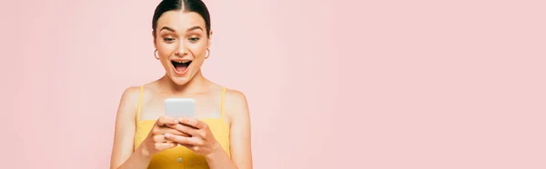 Choqué brunette jeune femme en utilisant smartphone isolé sur rose, panoramique shot — Photo de stock