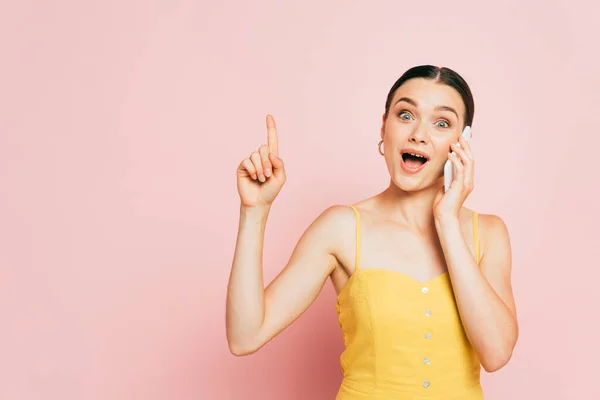 Excited brunette young woman showing idea gesture and talking on smartphone on pink — Stock Photo