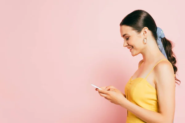 Seitenansicht der brünetten jungen Frau mit Smartphone isoliert auf rosa — Stockfoto