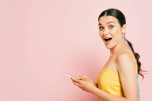 На вид здивованої брюнетки молода жінка, яка користується смартфоном ізольований на рожевому — стокове фото