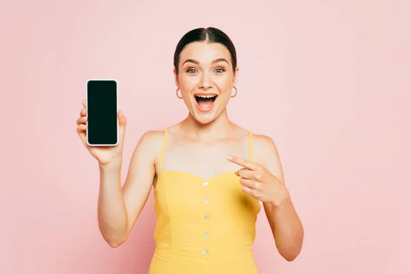 Mujer morena excitada apuntando a teléfono inteligente con pantalla en blanco aislado en rosa - foto de stock