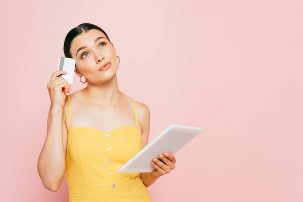Nachdenkliche brünette junge Frau mit digitalem Tablet und Kreditkarte isoliert auf rosa — Stockfoto
