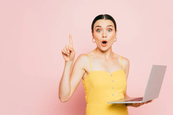 Surprised brunette young woman using laptop and showing idea gesture on pink — Stock Photo