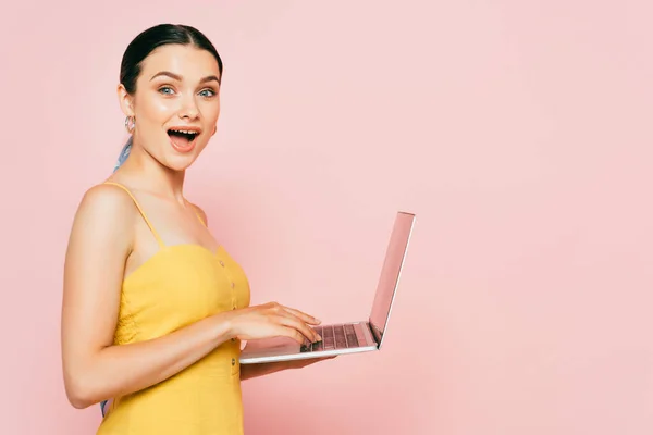 Vista lateral de la joven morena conmocionada usando el ordenador portátil en rosa - foto de stock