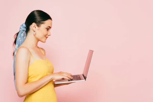 Vue latérale de la jeune femme brune en utilisant un ordinateur portable isolé sur rose — Photo de stock