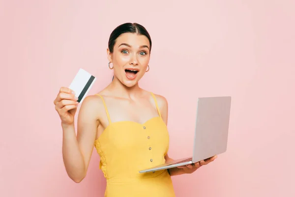 Возбужденная брюнетка молодая женщина держит кредитную карту и ноутбук на розовый — стоковое фото