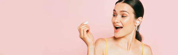 Excitada joven morena mirando auriculares aislados en rosa, tiro panorámico - foto de stock