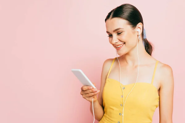 Morena mujer joven escuchando música en los auriculares y sosteniendo teléfono inteligente aislado en rosa - foto de stock