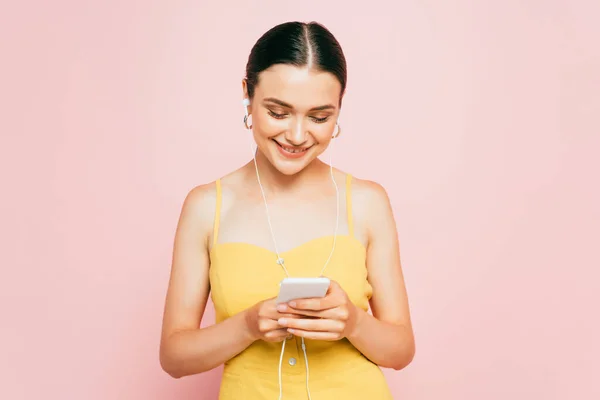 Jovem morena ouvindo música em fones de ouvido e segurando smartphone isolado em rosa — Fotografia de Stock