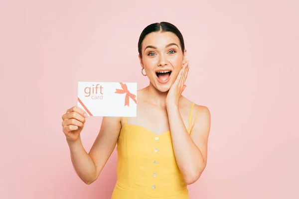 Excitada morena joven sosteniendo tarjeta de regalo aislada en rosa - foto de stock
